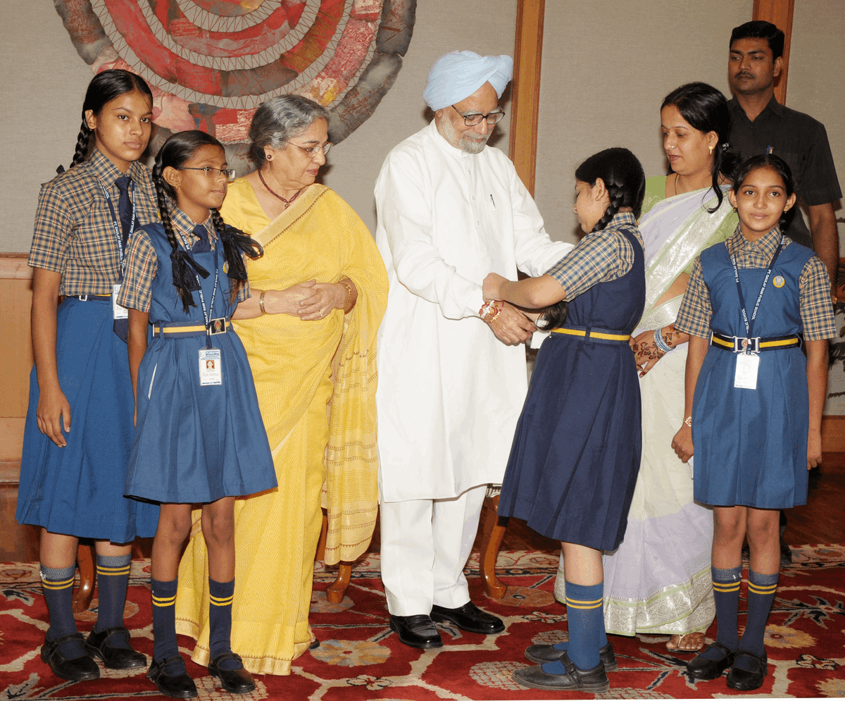 Crianças em idade escolar amarram rakhi no pulso do primeiro-ministro da Índia, Dr. Manmohan Singh, durante o Raksha Bandhan em Nova Délhi, em 2011, com Gursharan Kaur presente